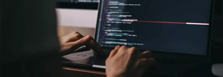 Close-up of hands typing code on a laptop in a dimly lit room.