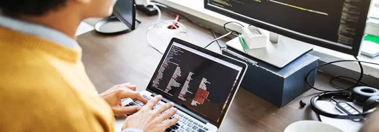 Person working on a laptop with code on the screen, seated at a desk with a large monitor.