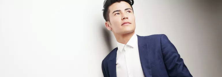 Man in a blue suit leans against a white wall, looking upwards.