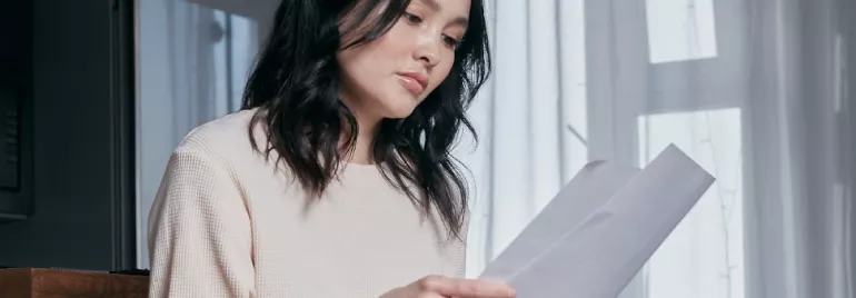 Woman with dark hair reads a letter by a window with soft light.