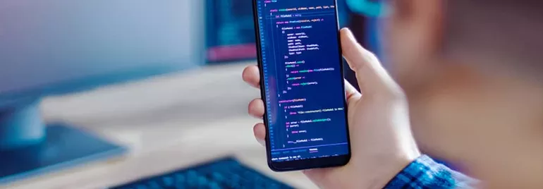 Person holding a smartphone displaying code, with a keyboard and monitor in the background.