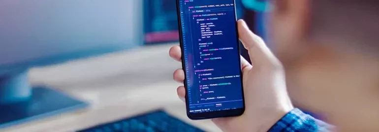 Person holding a smartphone displaying code, with a keyboard and monitor in the background.