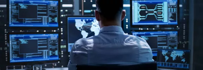 Person working at a desk with multiple monitors displaying data in a server room.