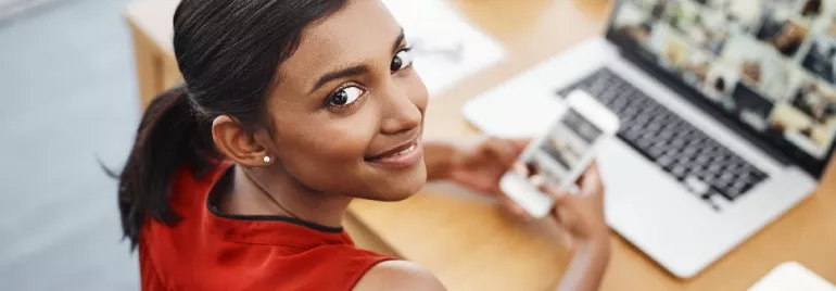Woman in red top using smartphone and laptop at desk, smiling.