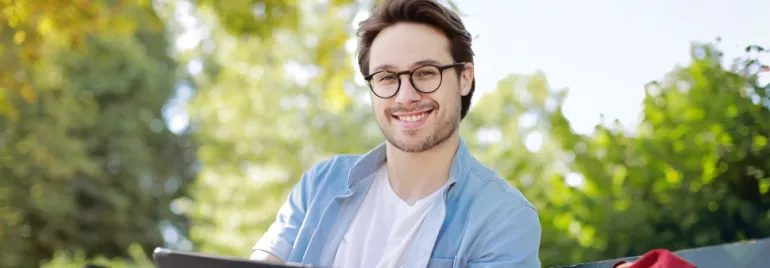 Man with glasses smiling outdoors, sitting on a bench.