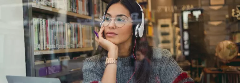 Woman with headphones studies at a library, gazing pensively outside.
