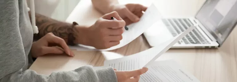 Two people reviewing documents at a desk with a laptop open.