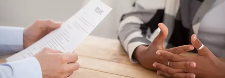 Close-up of an interview, one person holding a resume, another with folded hands on a wooden table.