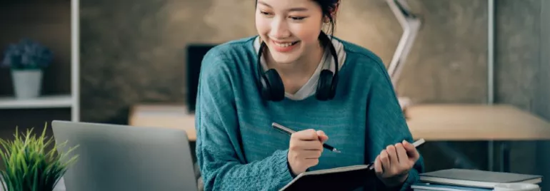 Woman with headphones studying at a desk with a laptop and notebook.