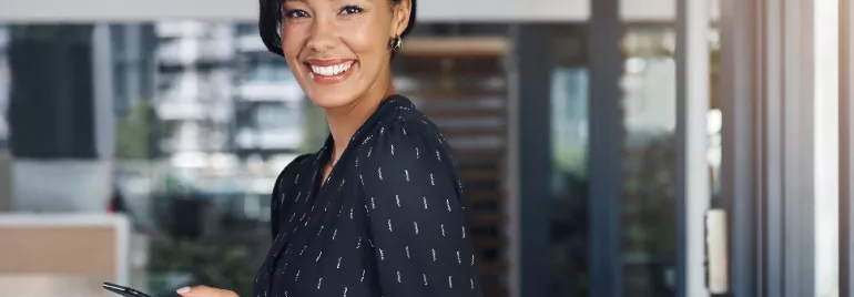 Woman in a black dress smiling and holding a phone in an office setting.