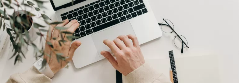 Person typing on a laptop with a notebook, pen, and glasses on a white desk.