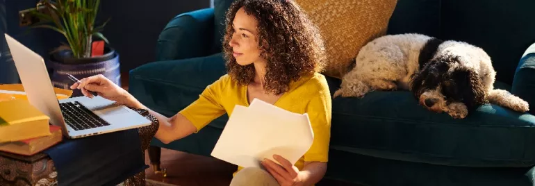 Woman working on a laptop with papers, sitting on the floor near a couch.