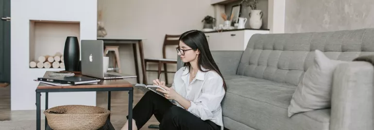 Woman sitting on floor by a coffee table with a laptop and notebook.