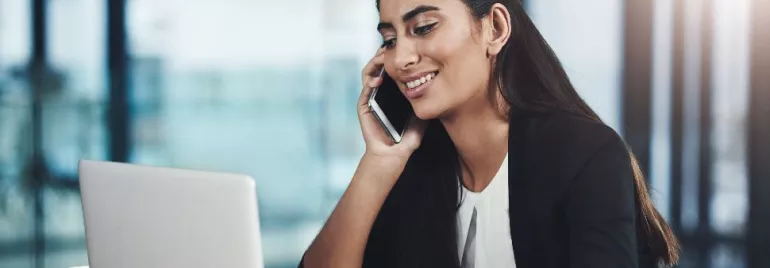 Woman smiling while talking on a phone and using a laptop.