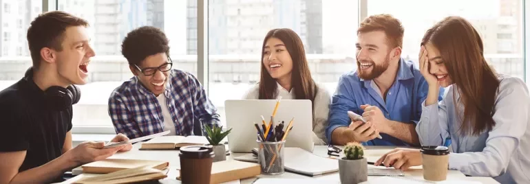 Group of young adults laughing and working together at a table with books, papers, and laptops.
