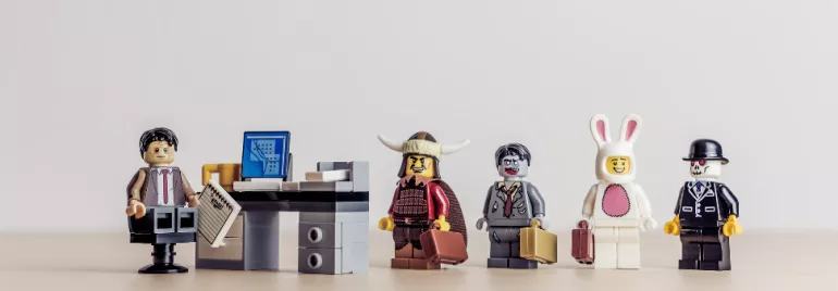 LEGO figures in costumes stand in a line, one sits at a desk with a paper, depicting a quirky office scene.