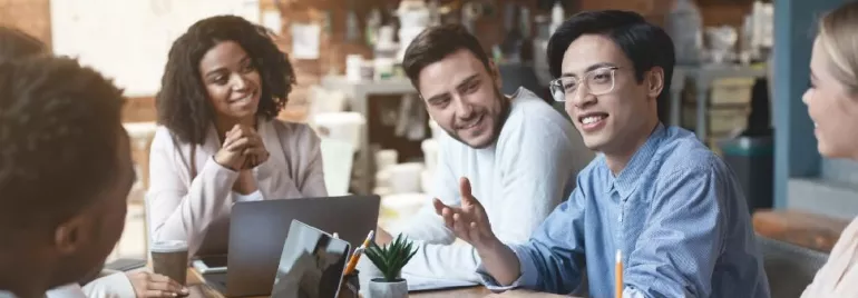 Diverse group of people in a casual meeting, discussing and smiling around a table with laptops.