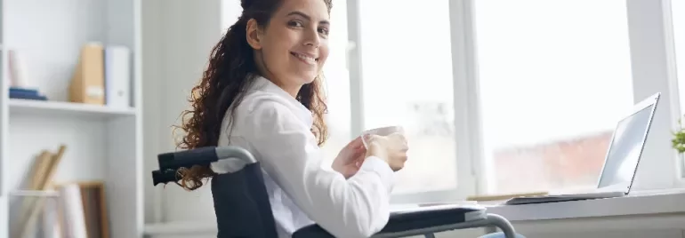 Woman in wheelchair smiling, holding a coffee cup next to a laptop.