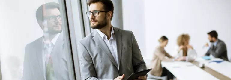 Businessman holding a tablet looks out the window; blurred meeting in the background.