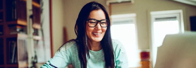 A woman with glasses smiling while working on a laptop at home.