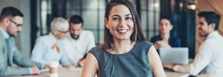 Smiling woman in a gray dress in an office setting with colleagues in the background.