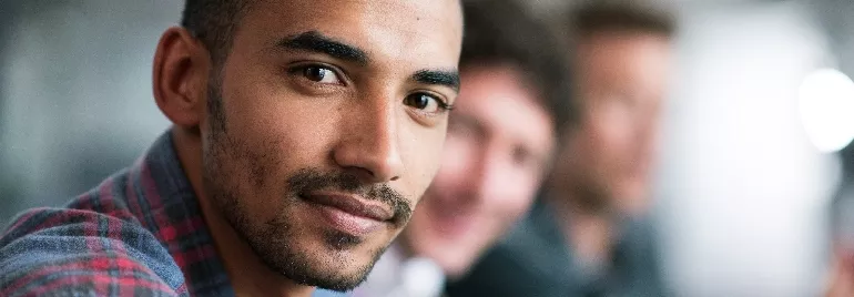 Smiling man in a plaid shirt, two people blurred in the background.