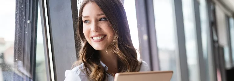 Woman smiling while holding a tablet by a window.