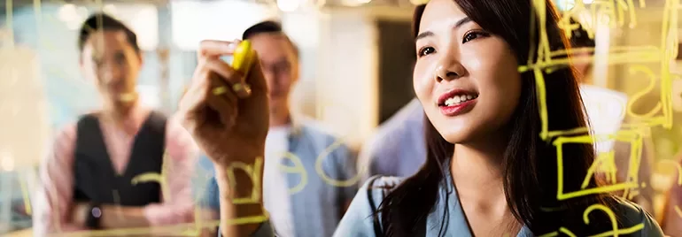 Woman writing on a glass board with colleagues in the background.