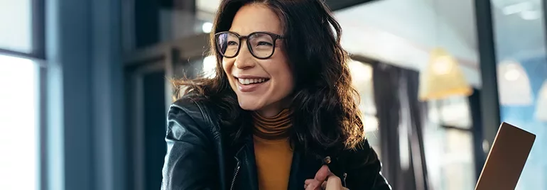 A woman in glasses smiling, sitting indoors with natural light.
