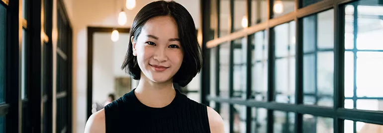 A smiling person stands in a modern hallway with glass walls.