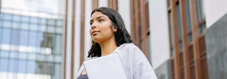 Person holding a laptop outdoors, standing in front of a modern building.
