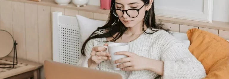 Woman wearing glasses, holding a mug and looking at a laptop.