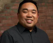 Smiling person in a dark shirt standing against a brick wall.