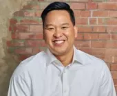 Man smiling in front of a brick wall, wearing a white shirt.