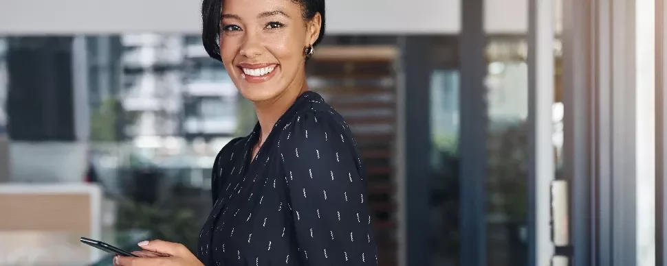 Woman in a black dress smiling and holding a phone in an office setting.