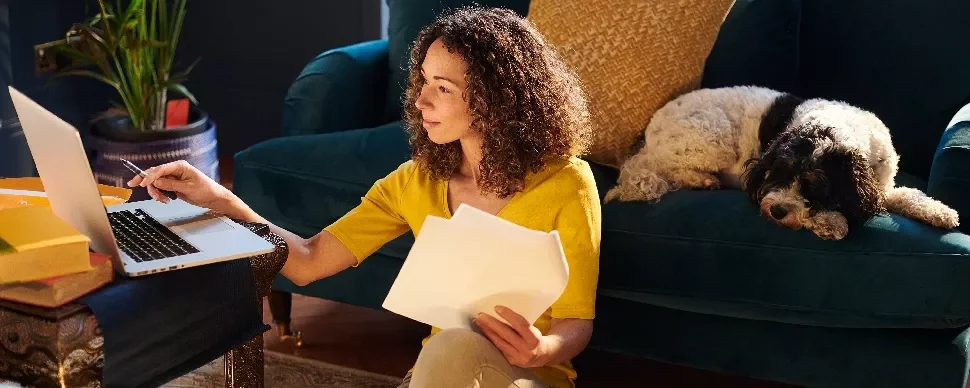 Woman working on a laptop with papers, sitting on the floor near a couch.