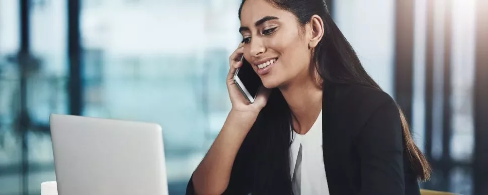 Woman smiling while talking on a phone and using a laptop.