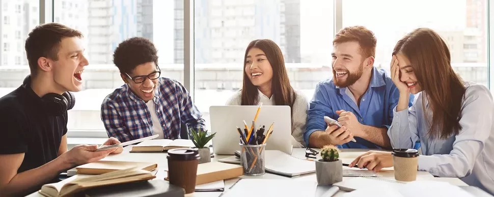 Group of young adults laughing and working together at a table with books, papers, and laptops.