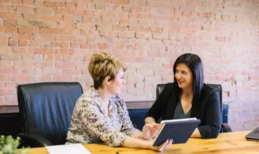 Two women discussing work at a conference table, one holding a tablet.