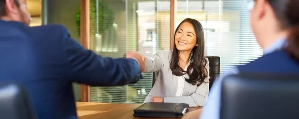 A woman smiles while shaking hands with a person across a table in an office setting.