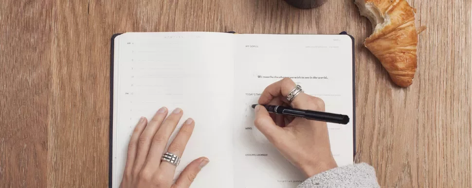 Person writing in a notebook on a wooden table with a croissant and coffee nearby.