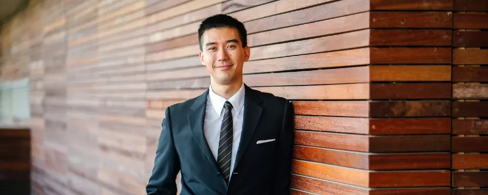 Man in a suit leaning against a wooden wall, looking at the camera.