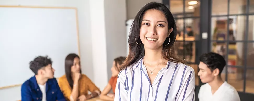Smiling woman in a striped shirt stands in front of a group of people in a modern office environment.