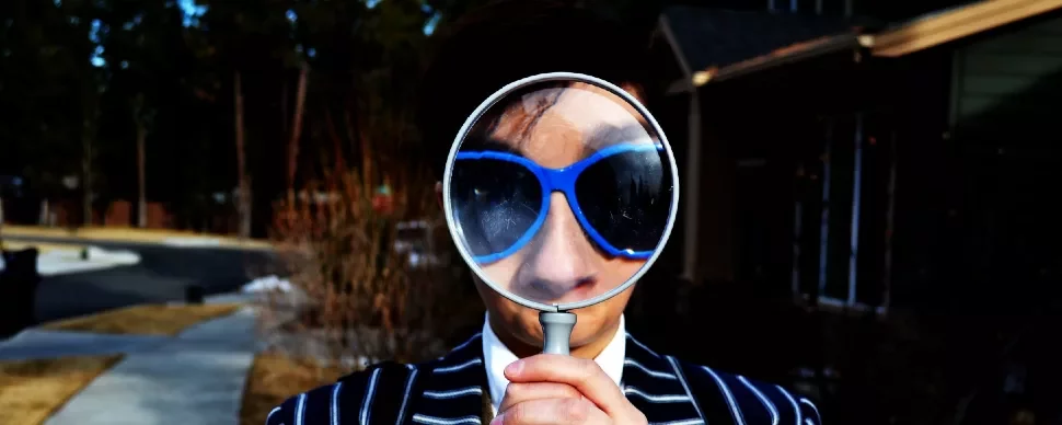 Person in blue sunglasses holding magnifying glass, outside on a sidewalk.