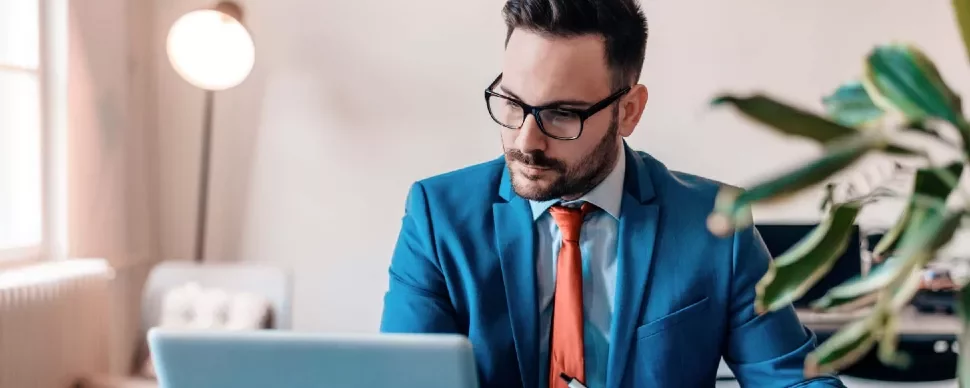 Man in a blue suit and red tie working on a laptop in an office.