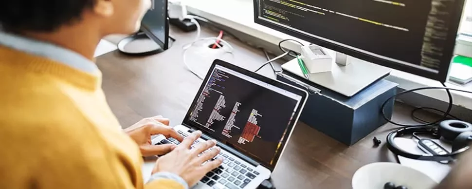 Person working on a laptop with code on the screen, seated at a desk with a large monitor.