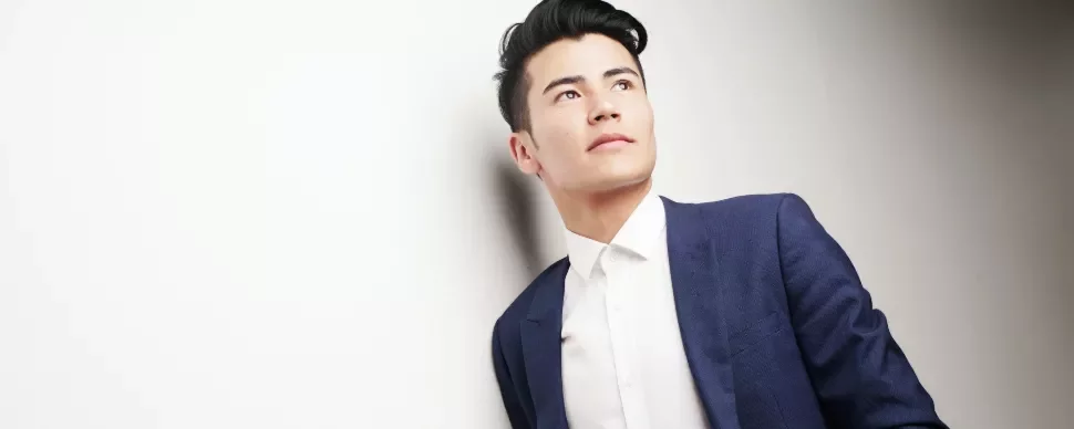 Man in a blue suit leans against a white wall, looking upwards.