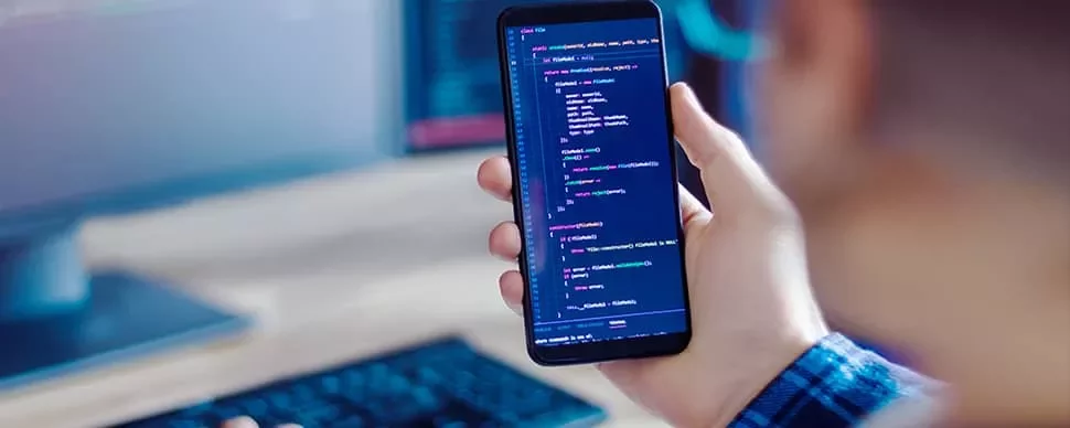 Person holding a smartphone displaying code, with a keyboard and monitor in the background.