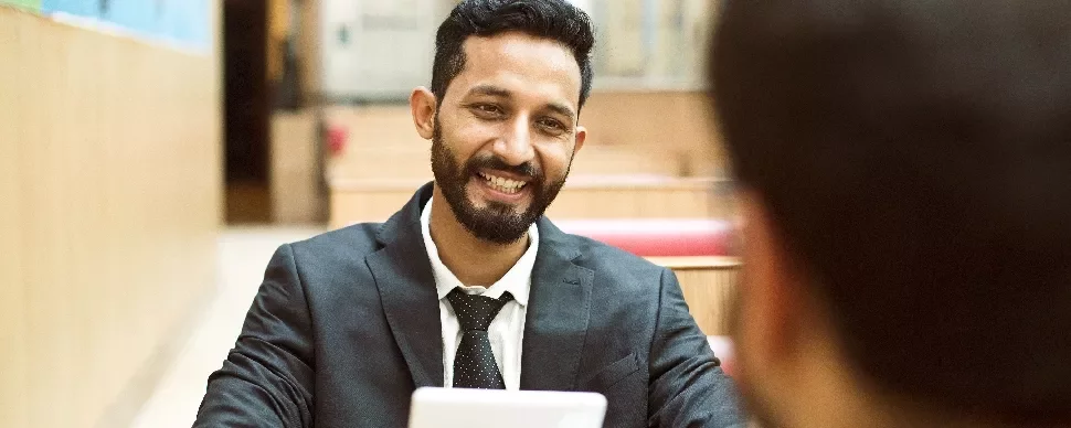 Smiling man in a suit holding a tablet, sitting at a table.