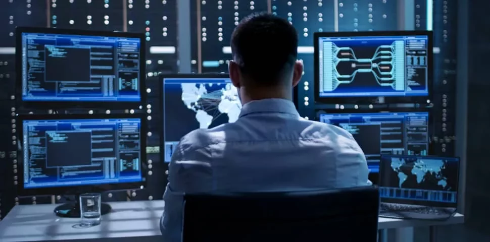 Person working at a desk with multiple monitors displaying data in a server room.